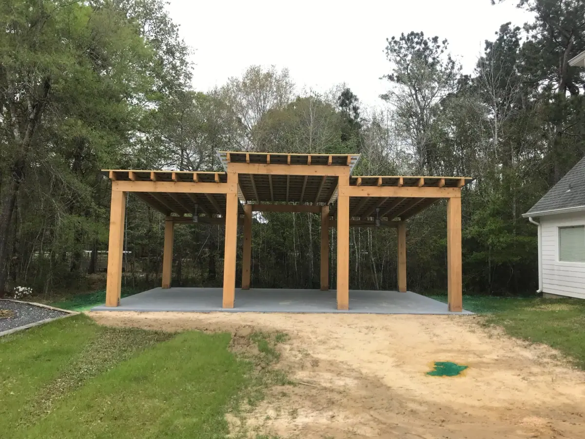 Open timber pavilion frame on a concrete slab in a wooded yard