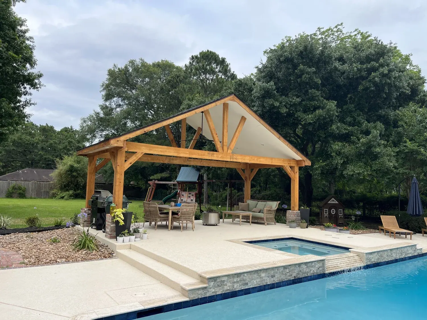 Heavy-timber gable pavilion beside a pool: treated-pine trusses and posts, dark metal roof, outdoor furniture underneath